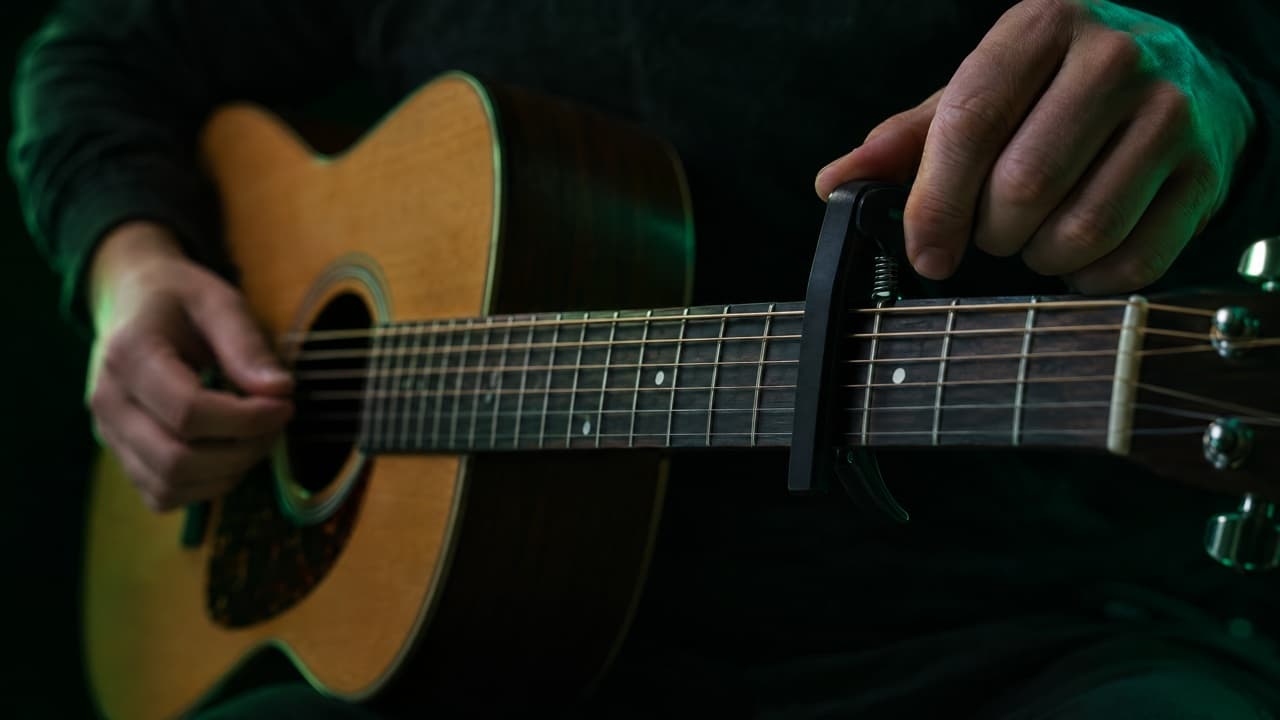 Hand setzt einen schwarzen Federkapodaster hinter einem Bund auf den Hals einer Akustikgitarre